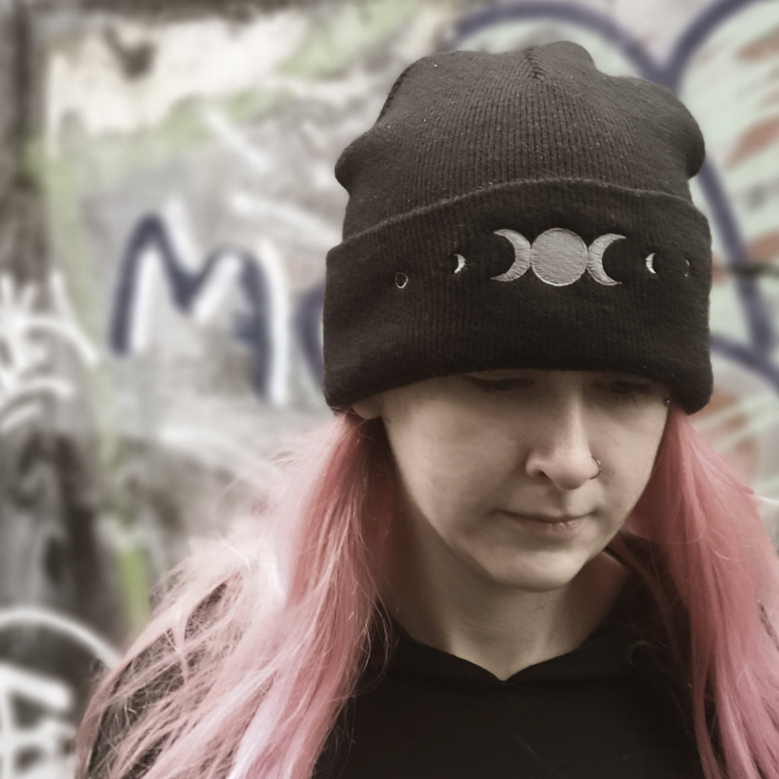 Person wearing a black beanie with moon phases in front of graffiti.