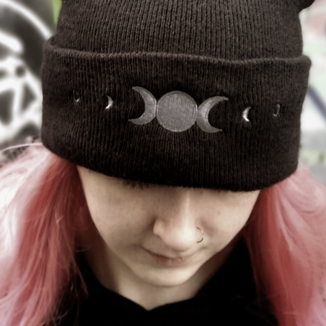 Person wearing a black knit beanie with moon phase design, pink hair, and red jacket.