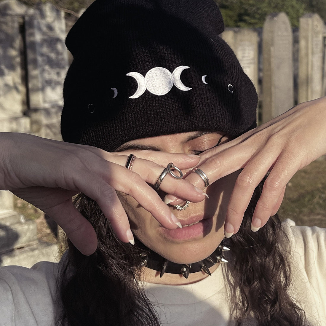 Person wearing a black beanie with moon symbols, holding hands over face, in front of stone wall.